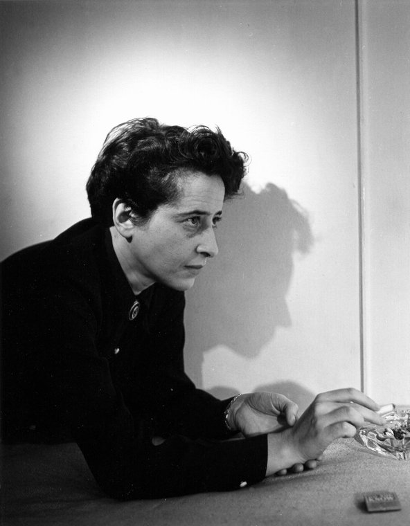 Portrait of German-born American political theorist and author Hannah Arendt (1906 - 1975) in profile, a cigarette in hand, as she leans across a table. Arendt is dressed in black and her dark hair is cut short. She stares across the table at something out of frame. Her face is silhouetted in profile on the unadorned wall beside her.