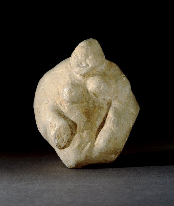 Carved chalk female figurine, anaively excecuted. Lumpy and lopsided in appearance. Finished with a flint blade, leaving striations. The formless head has eyes, nose and mouth crudely rendered. There is no neck, the front of the head being separated from the body by a groove. The breasts are lumpish, with nipples perhaps indicated by slight pits, as is the navel. Two crude arms rest on a protuberant belly. Possible modern forgery?