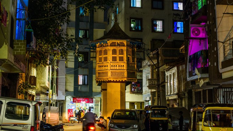 Night scene of a busy street in an urban area with a distinctive traditional architectural structure in the center, surrounded by modern buildings and lit shops.