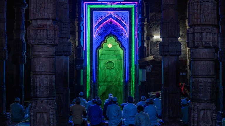 People participating in prayer in a mosque, which features vibrant blue and green light projections creating an illuminated archway over the mihrab.