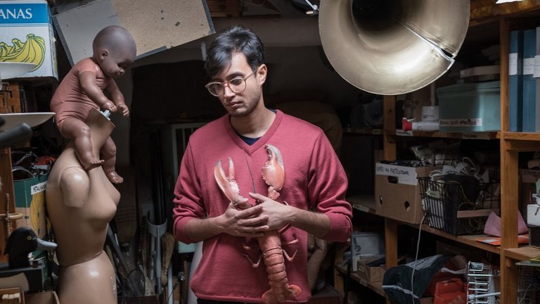 An individual in a cluttered workshop holds a large model of a lobster, surrounded by various objects including a mannequin.