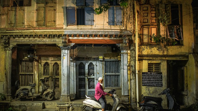 An individual rides a scooter in front of a row of colourful, time-worn buildings in an urban setting during sunset.