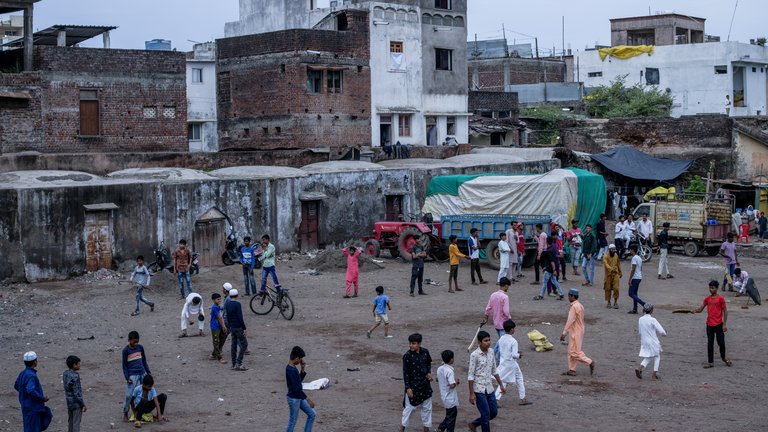 Children and adults playing and walking in an open area surrounded by old buildings and vehicles in a busy community setting.