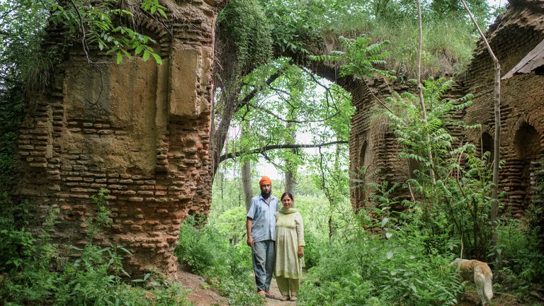 Two people standing under the arches of a dilapidated brick structure, surrounded by lush green foliage.