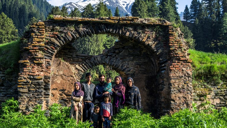 A family of six standing in front of an ancient stone arch with lush greenery and distant snow-capped mountains.