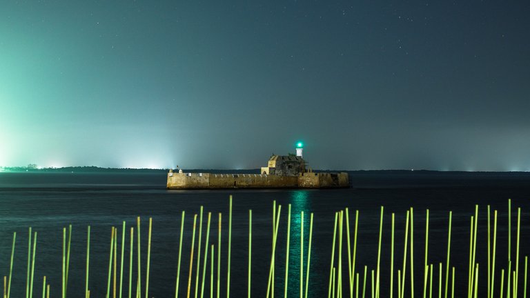Night view of Fort Boyard with illuminated beacon, surrounded by sea, under a clear sky with glowing horizon lights in the distance.