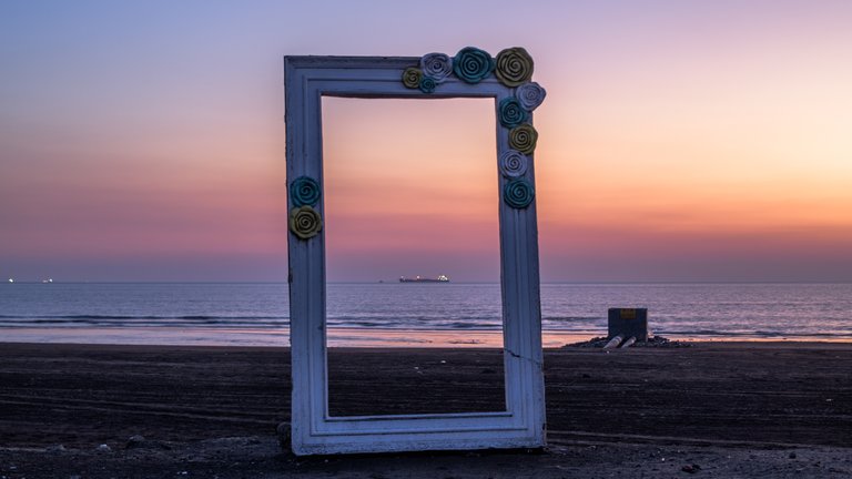 Decorative frame with fabric flowers on top positioned on a beach, framing a view of the ocean under a colourful sunset sky.