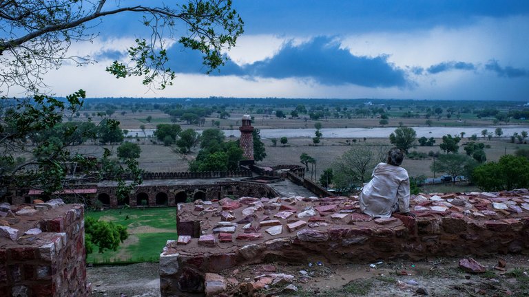 Person sitting on a brick wall, gazing at a vast landscape with dark clouds overhead, near the historic Fatehpur Sikri in India.