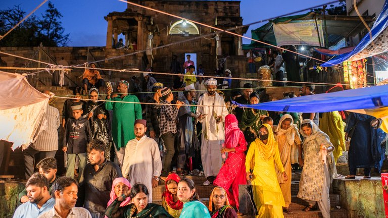 Group of people gathered at an outdoor event near a historic structure at dusk, with some wearing traditional attire.