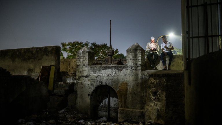 Two people are sitting atop an old wall, engaging in conversation under moonlight, surrounded by rustic surroundings.