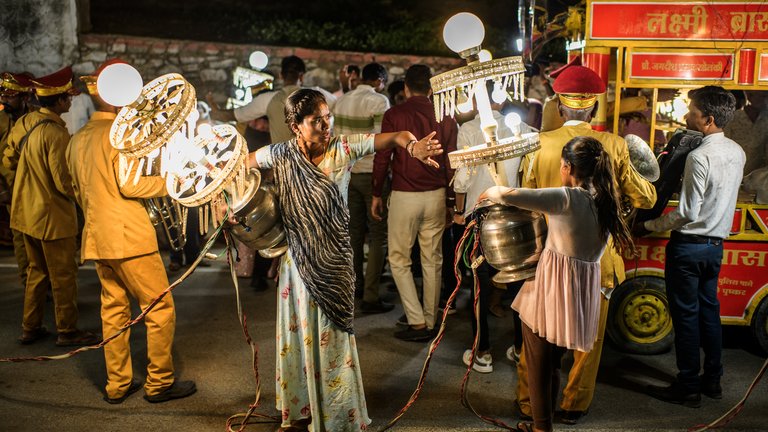 During a wedding procession, a woman in traditional attire interacts with another individual carrying a large pot amidst street vendors.