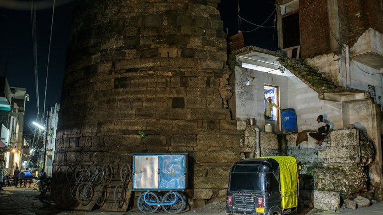 Nighttime scene in a bustling urban area with a historic stone structure, people interacting near small shops, and various transport modes including a tuk-tuk and a bike cart.