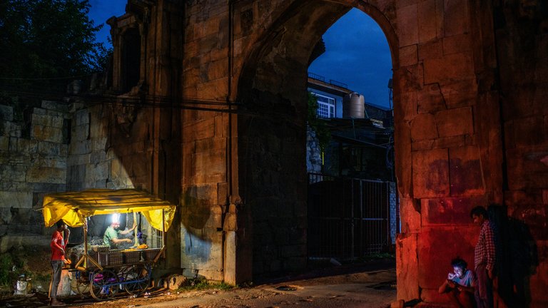 Nighttime scene at an old arched gateway where a person operates a food cart under a shelter, illuminated by lamps, while another person sits nearby using a phone.