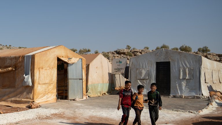 Three children walking toward a group a group of tents on the first day of school in a refugee camp in the countryside of Idlib, on 9 October 2021