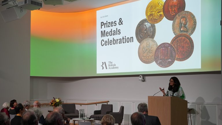 An audience listens attentively in a modern auditorium as a woman speaks at a podium during "The British Academy Prizes & Medals Celebration," displayed on a large screen.