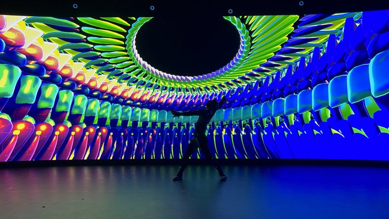 A person in silhouette dancing in front of a swirling, multicoloured neon light display generated by a LED screen that wraps around the walls and ceiling of the space.