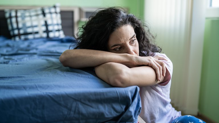 Worried woman sitting on floor next to bed.