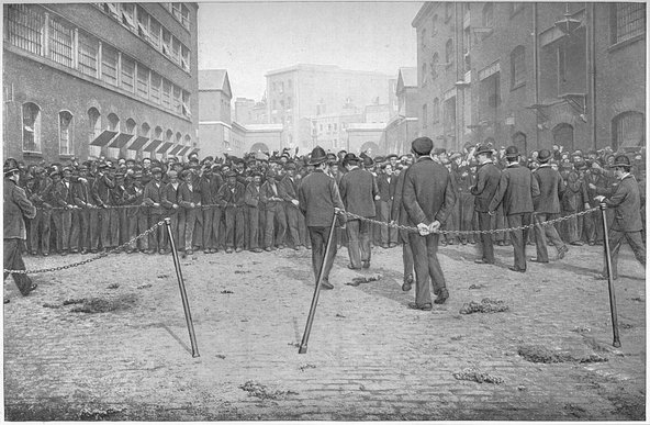 Black and white illustration of a crowd of men gathering by the fence at the London Docks surrounded by buildings.