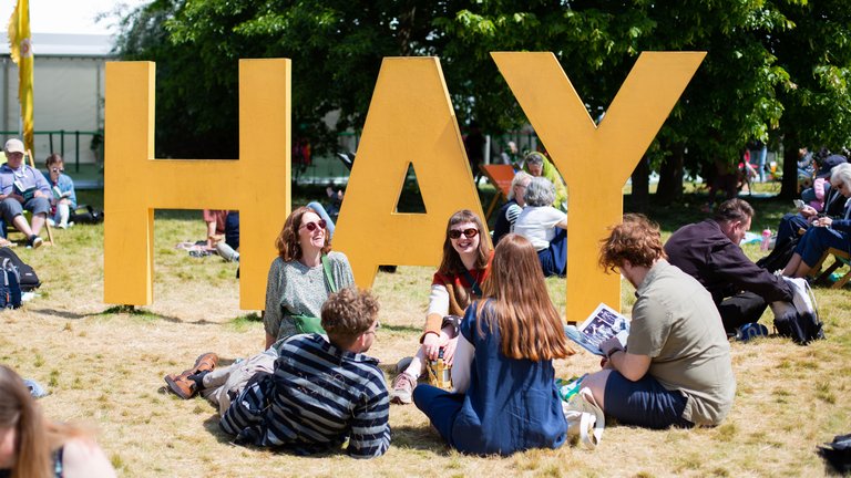 People sitting on the grass in front of a 3D sign saying 'HAY'