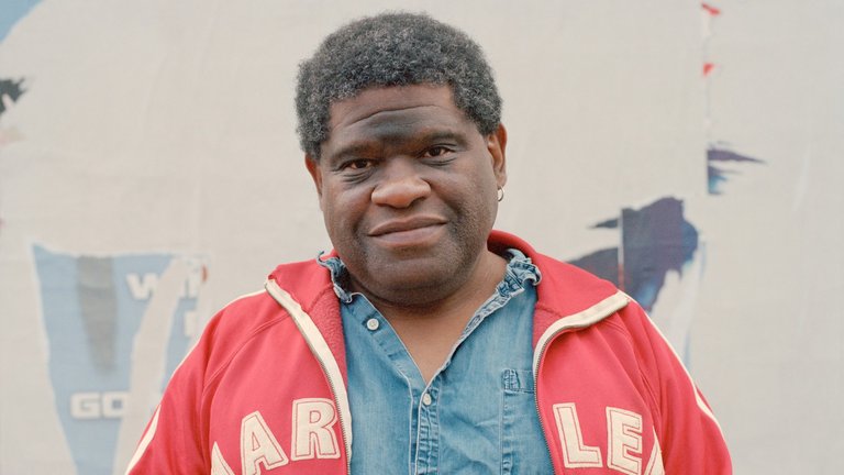 An image of Gary Younge standing against a wall wearing a red jacket.