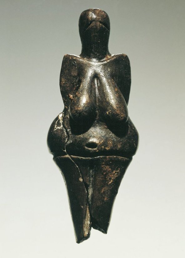 A dark, clay and bone powder female figurine. The head has eyes and mouth crudely rendered. A neck joins roughly symmetrical shoulders and arms. The figure's breasts, hips and belly button are prominently represented. There is a crack running down the lower part of the torso and right leg.