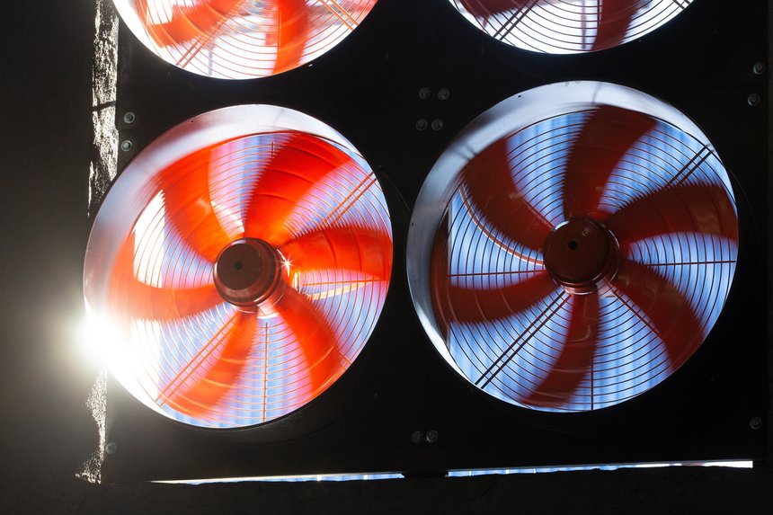 Four orange industrial cooling fans in operation at a cryptocurrency farm. Sunlight shines through two of the vents.
