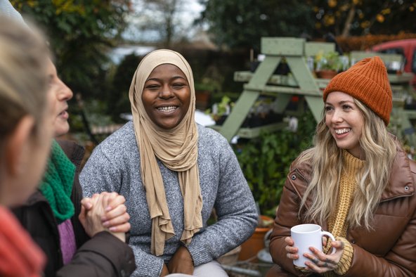 Four friends in warm outdoor clothing sit together in conversation in a garden or allotment.