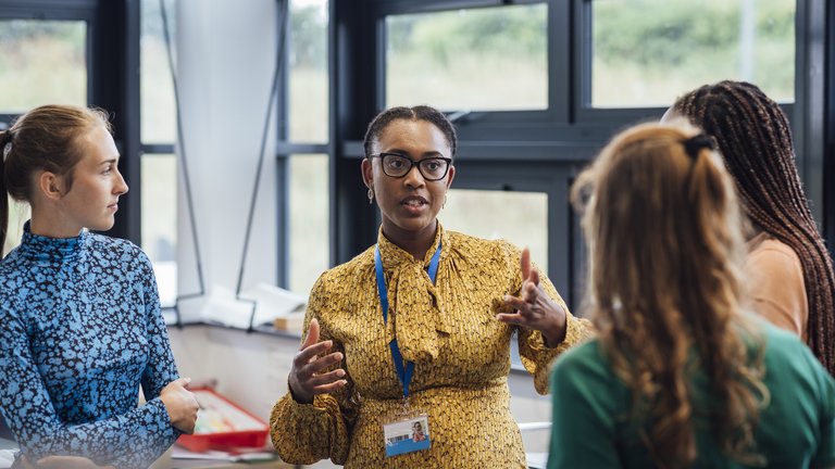 A shot of a mid-adult secondary school teacher talking with her sixth form students in class, they are wearing casual clothing and discussing what they learned in class