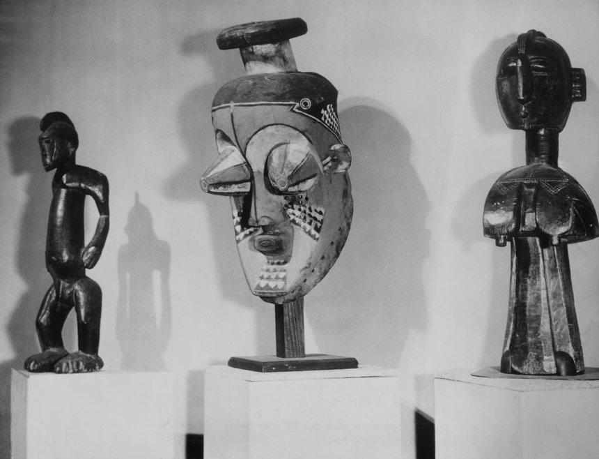Black and white photograph of three wooden artworks on pedestals in front of a plain background. On the left is a dark wooden figure representing a man. In the centre is a stylised facemask with protruding eyes and patterned facepaint. On the right is a dark wooden fertility symbol representing a woman, with a head on a long, thin neck, and prominent breasts.