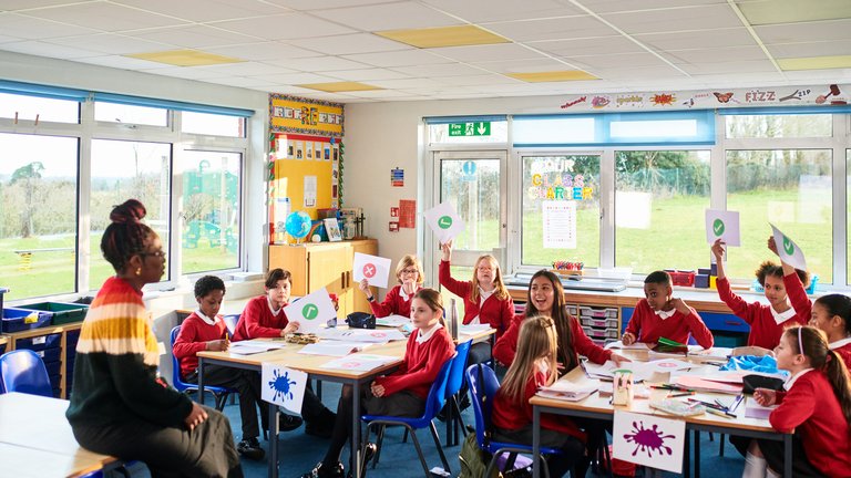 Teacher talking to class of primary school children holding up pieces of paper