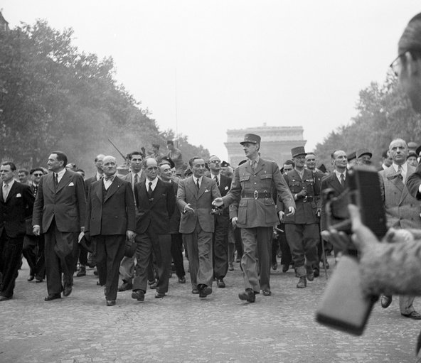 General Charles de Gaulle leads a procession down Champs-Elysees as part of the celebration of the liberation of Paris. The people in the photograph are all men, some in civilian business suits and others in military uniform. The top of the Arc de Triomphe is visible in the background.