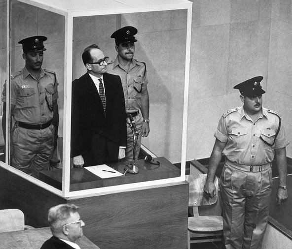 Adolf Eichmann, flanked by two guards, stands in a dock shielded by bullet proof glass while on trial. Eichmann wears a dark suit and thick-rimmed glasses. His defence attorney, Robert Servatius, is in the foreground of the photo, facing away from the camera.