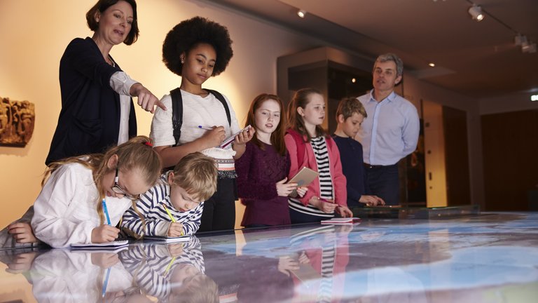 Two adults and a group of young students examining an interactive display in a museum.
