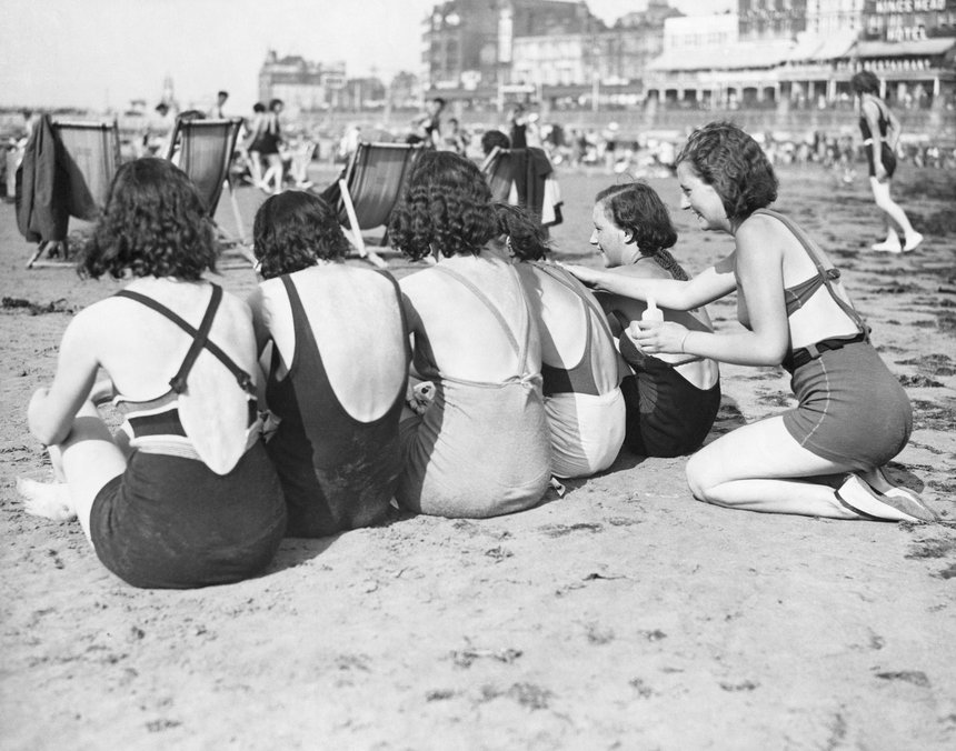 Black and white photo of a group of women friends in bathing suits on the beach at Margate, 1934. Five friends sit in a row with their backs to the camera, while a sixth friend applies sun cream to one of the group.