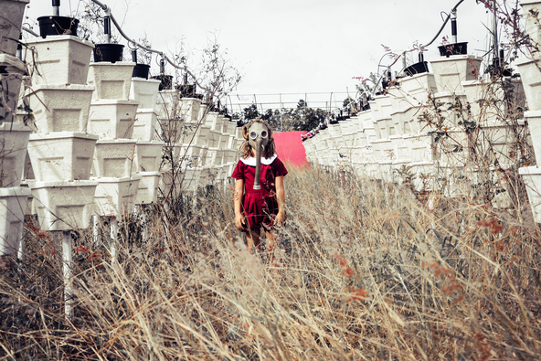 A young child in a red dress with a white collar stands in an overgrown field between two rows of stacked white planters, wearing an old-fashioned gas mask.