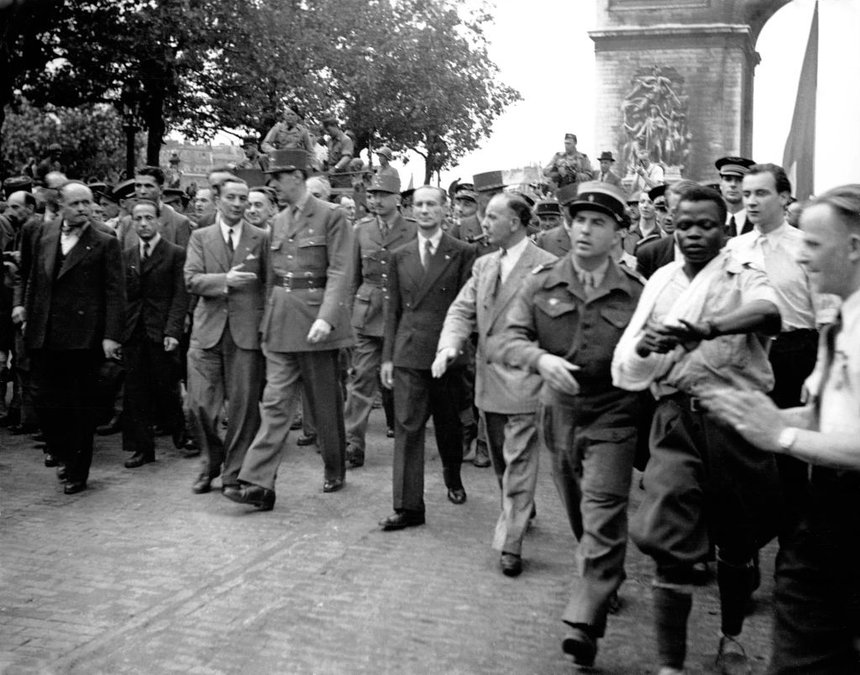 Georges Dukson, a Black soldier wearing a white shirt and culottes, with his arm in a sling, is manhandled by a white official during the parade led by Charles de Gaulle to mark the Liberation of Paris, 1944. De Gaulle, wearing a military dress uniform, walks at the front of the parade a few meters from Dukson, at this point apparently having not noticed the altercation taking place.