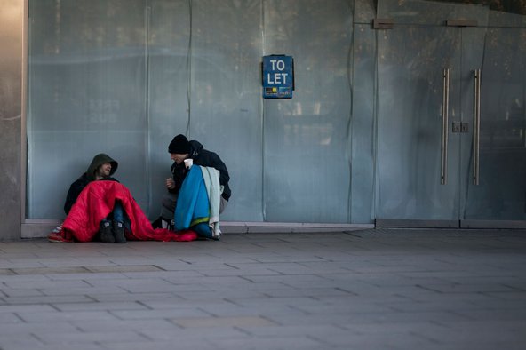 Two men sitting on the ground by an empty glass building unit with a sign saying "To let". They're covering themselves with blankets and one of them is holding a plastic cup for change.