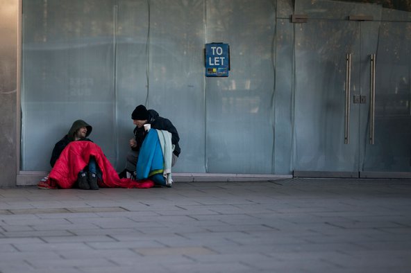 Two men sitting on the ground by an empty glass building unit with a sign saying "To let". They&#x27;re covering themselves with blankets and one of them is holding a plastic cup for change.