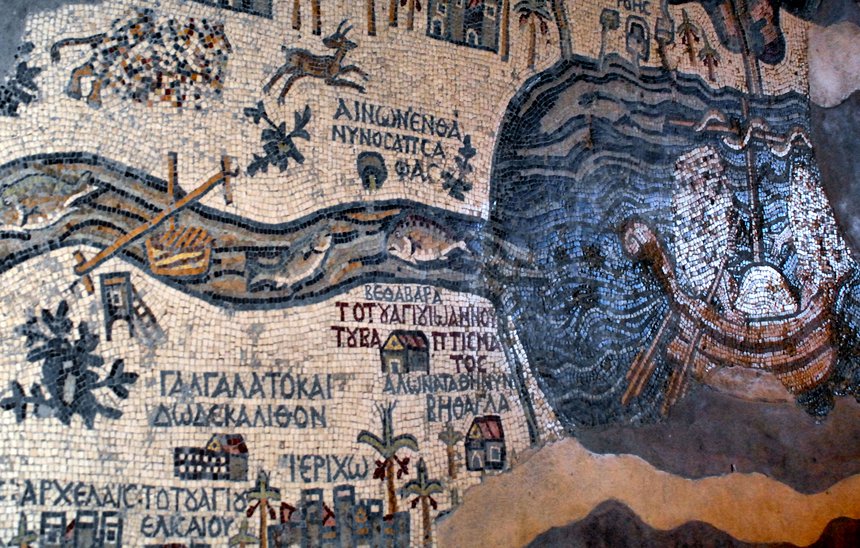 Colourful photograph of a wall mosaic, which illustrates a fragmented map of the Holy Land, including the blue sea with a ship on the right side and a river leading into the white landmass that makes up the entirety of the image with animals, houses, trees and villages spread out on it. Labels in blue mark the names of locations and landmarks on the map.