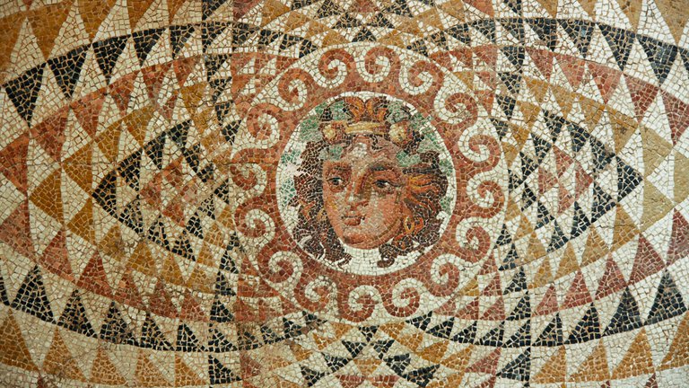 A colourful mosaic with the face of the Roman god Dionysus depicted in the centre, wearing a vine wreath and surrounded by a geometric pattern.