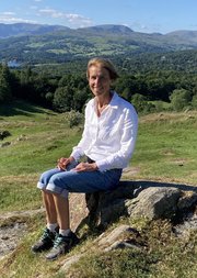 Sarah Nettleton sitting on rock against a backdrop of green hills, trees and a lake in the top left