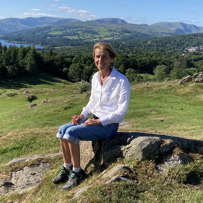 Sarah Nettleton sitting on rock against a backdrop of green hills, trees and a lake in the top left