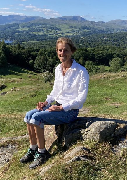 Sarah Nettleton sitting on rock against a backdrop of green hills, trees and a lake in the top left