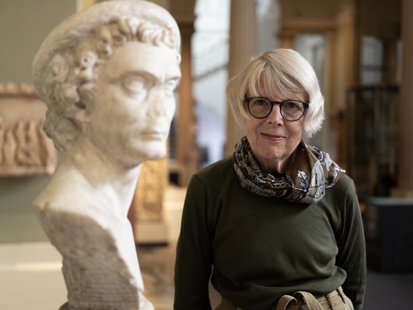 Susan Walker standing in a museum beside a marble bust of a man with curly hair and wearing a hat tilted to one side. The bust's nose is missing.