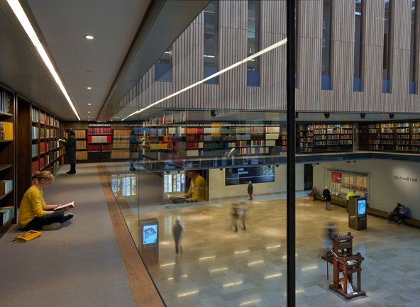 Colourful photograph of the library showing two levels, the ground floor with blurry figures walking around and the upper floor overlooking it through a glass wall, where a young woman with a bright yellow shirt is sitting down and reading a book on the ground. The upper floor is filled with colourful bookshelves.