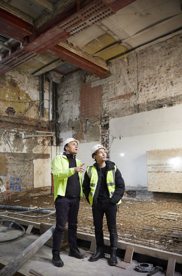 A member of the Wright & Wright team and a member of the construction team discuss the project while standing in on of the new spaces which is in the early stages of renovation, with exposed brick work and metal beams visible in the background. Both colleagues wear hard hats and high-visibility clothing.