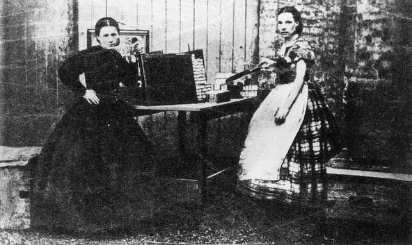 An old, black-and-white photograph of two women in long dresses standing by a table with matchsticks.